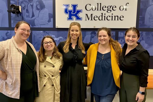 five CELT staff posing and smiling in front of a wall with a blue banner installation and the UK College of Medicine lockup prominently displayed