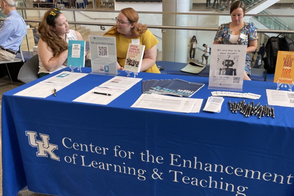 three women behind a table with a blue cloth on which "UK center for the enhancement of learning and teaching" is printed, with papers and books on top of the table