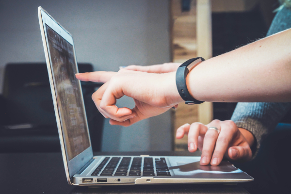 a hand points at a screen of an open laptop while another pair of hands types on the keyboard
