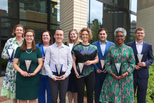 nine people stand holding statuettes in front of an academic building