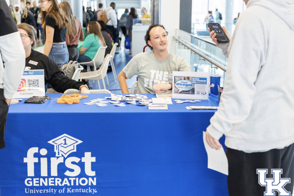 a student with a first gen t shirt sits behind a table with a first gen blue table skirt at a student event in the student center