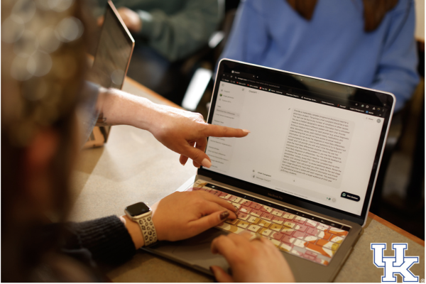 the camera frame peers over someone's shoulder as they use ChatGPT on a laptop and someone else out of frame points to the text exchange on the screen