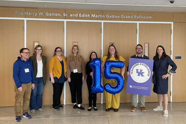 eight people stand smiling behind a large balloon "15" and a sign with the UK teaching excellence symposium logo