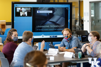 four faculty discussing around a table in front of a projection screen with a "leveraging technology" slide displayed