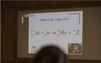 Picture of a slide in the background with an equation on it. The top of an out of focus head is in the foreground