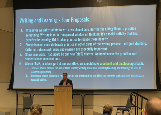 presenter on a stage with a powerpoint slide behind him titled "writing and learning: four proposals"