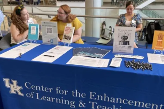 three women sit behind a table with a blue cloth with the CELT logo and handouts on the table at new faculty orientation 2023 in the social staircase atrium of the gatton student center
