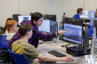 students sit in a row of desks with computer screens in front of them with one student standing and pointing at the screen of another student's monitor showing a CAD-like interface