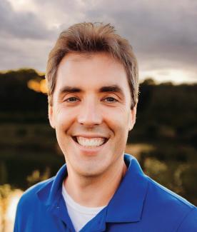 a man smiling against a sunset treeline background in a close up headshot