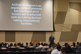 a man in a dark suit holds a microphone and speaks to a seated audience in a ballroom with a large slide projected about the definition of AI