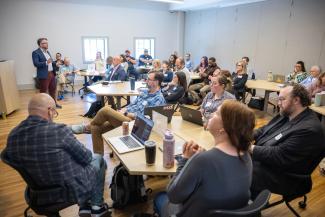 a man in a blue blazer stands in a wood-floored brightly lit room in front of an audience of about 30 people at small tables for an AI workshop