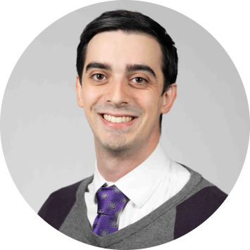 a man with short black hair smiling while wearing a white shirt and purple tie under a black and gray sweater vest