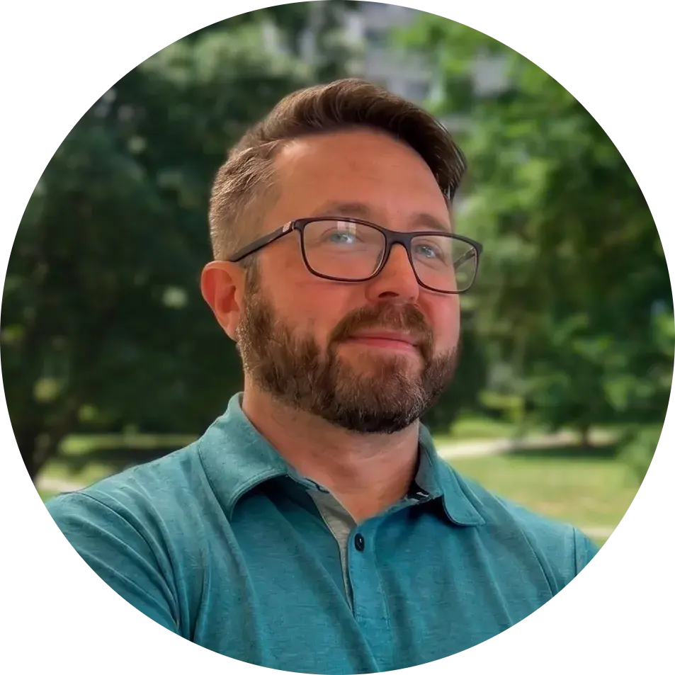 a man with short brown hair, glasses, and a beard smiles with green campus landscape in the background